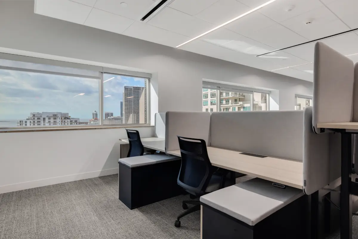 Modern open-plan office layout featuring modular workstations with light wood desktops, grey fabric privacy screens, and black ergonomic task chairs in a professional corporate workspace.